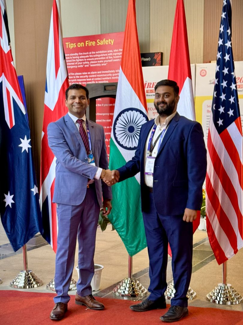 Two man shake hands in front of the flag of India.