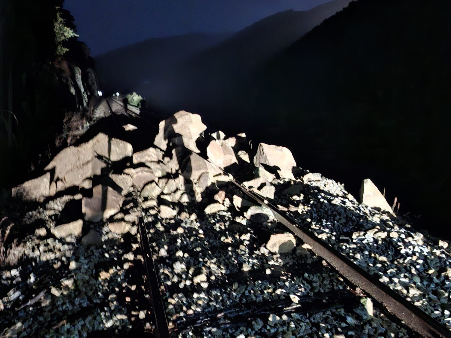 A large rockfall blocking a railway through the mountains. The photo is taken at night with the rockfall illuminated by torches/vehicle lights
